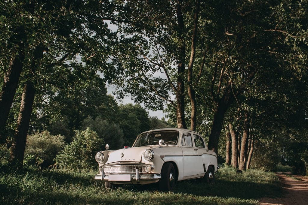 A white car on the side of a dirt road lined by trees